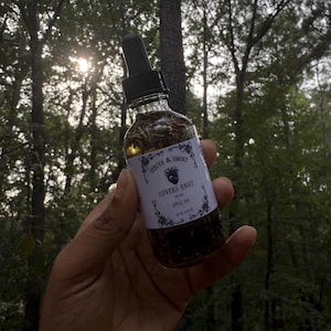 Könnte beinhalten: Eine klare Glasflasche mit einer schwarzen Pipette, gefüllt mit einem Lovers Knot Zauberöl. Die Flasche hat ein weißes Etikett mit schwarzem Text und floralen Akzenten. Das Öl enthält Kräuter.
