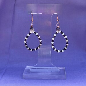 May include: A pair of teardrop-shaped earrings. Each earring features a copper-colored hook and is adorned with alternating black and white beads. The earrings are displayed on a clear acrylic stand against a purple backdrop.