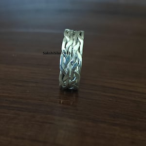 May include: A silver ring with a woven design. The ring features an intricate pattern of interwoven metal strands, creating a textured and detailed appearance. The ring is displayed on a wooden surface.