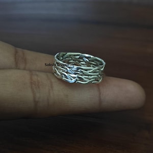 May include: A silver ring with a woven pattern. The ring is made of a shiny, metallic material and has a textured surface. The ring is displayed on a finger against a dark brown background. The text "SakshiSilverHub" is visible on the image.