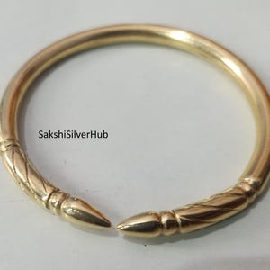 Peut inclure: Un bracelet jonc doré avec des extrémités décoratives. Le bracelet a une finition lisse et polie avec des détails gravés près des extrémités pointues. Le texte "SakshiSilverHub" est visible sur le bracelet.