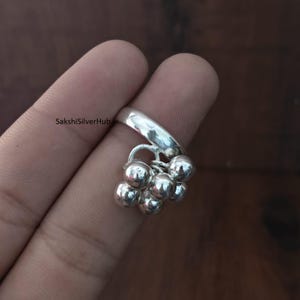 Puede incluir: Un anillo de plata con un grupo de pequeñas cuentas redondas. El anillo está sujeto por un dedo, y las cuentas cuelgan debajo de la banda. El anillo es brillante y reflectante, y las cuentas están agrupadas. Una joya.