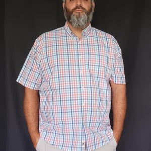 May include: A short-sleeved plaid shirt in red, white, and blue. The shirt has a button-down collar and is paired with light gray shorts. The shirt features a small logo on the bottom hem. The man is standing in front of a black background.