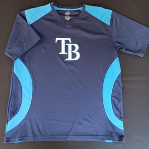 Pode incluir: Camiseta esportiva azul marinho com detalhes em azul claro e as letras "TB" em branco. A camiseta tem decote em V e mangas curtas. A camiseta é feita de um material respirável semelhante à malha.