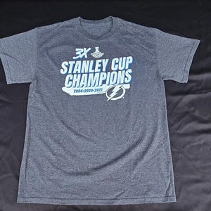 May include: Gray t-shirt featuring the text "3X STANLEY CUP CHAMPIONS" in white and blue, with the years 2004-2020-2021. The shirt also displays the Tampa Bay Lightning logo. The shirt is made of a soft material.