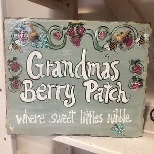 May include: Hand-painted sign with the words "Grandma's Berry Patch" in white script. The sign is light blue with decorative elements including bees, berries, and floral designs. The phrase "where sweet littles nibble" is also included.