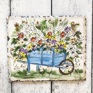 May include: A hand-painted wooden sign featuring a blue wagon overflowing with colorful flowers. The wagon has a wooden wheel and is set against a whitewashed wood background.