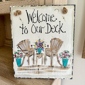 May include: Welcome to our deck sign with a painted image of two chairs, a table, and flowers in pots. The sign is made of slate and has a rustic look.