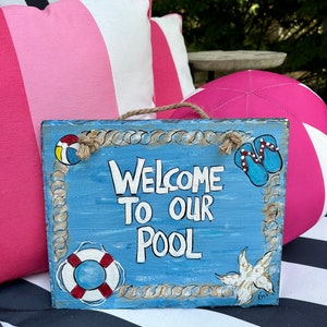 Puede incluir: Un letrero de madera azul con ribete de cuerda blanca y las palabras "Welcome to our Pool" pintadas en blanco. El letrero tiene un salvavidas rojo y blanco, una chancla azul y blanca y una estrella de mar blanca pintadas.