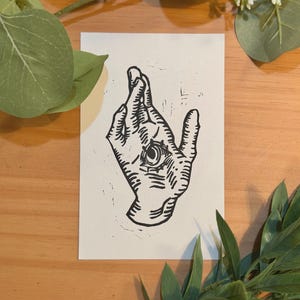May include: A rectangular white card featuring a black ink block print illustration of a hand with fingers crossed. The palm of the hand contains a detailed, centered eye design. The artwork uses bold, textured lines to define the hand's anatomy.