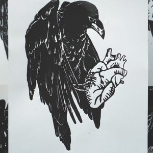 May include: Black and white linocut print of a raven holding a human heart. The raven is depicted with detailed wings and a slightly downward gaze. The heart is intricately detailed, with visible veins and chambers. The artwork is on a white background.