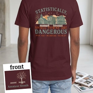 May include: A maroon t-shirt featuring a graphic of three houses behind a white picket fence with the text: Statistically, SMALL TOWNS ARE DANGEROUS POP. 412 MURDERS / YEAR: 37. A small inset shows the front design with a tree and the text: Amateur Sleuth.