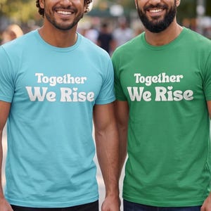 May include: Two short-sleeved crew neck t-shirts, one in light blue and one in green, both featuring the white text Together We Rise printed on the chest. The shirts have a standard fit with a round neckline and short sleeves, made of solid fabric.