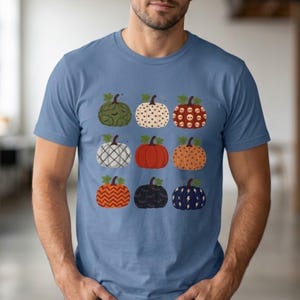 May include: A blue short-sleeve t-shirt featuring a graphic print of nine pumpkins arranged in a three-by-three grid. Each pumpkin displays a distinct pattern, including skulls, polka dots, lightning bolts, chevron stripes, and plaid designs.