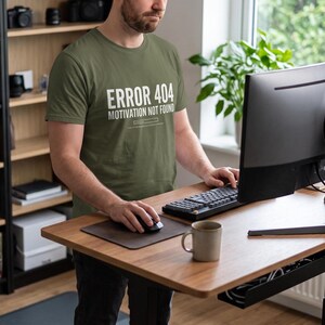 May include: An olive green short-sleeved t-shirt featuring the white text ERROR 404 MOTIVATION NOT FOUND with a loading bar graphic underneath. The shirt has a crew neckline and a standard fit, made of a solid-coloured fabric.