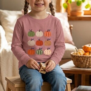 Peut inclure: Un t-shirt à manches longues rose poudré orné d'une grille de neuf citrouilles aux motifs et couleurs variés, incluant du vert, de l'orange, du blanc et du beige avec des feuilles. Porté avec un jean bleu et un petit bloc en bois.