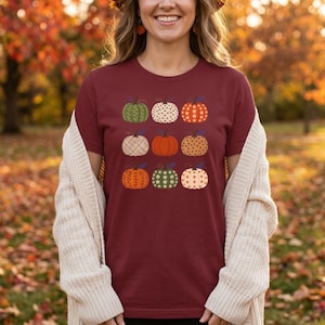 Peut inclure: Un t-shirt à manches courtes bordeaux orné d'une grille de neuf citrouilles aux motifs et couleurs variés, incluant du vert, de l'orange et du crème avec des pois, des rayures et des fleurs, porté sous un gilet en maille épaisse crème.