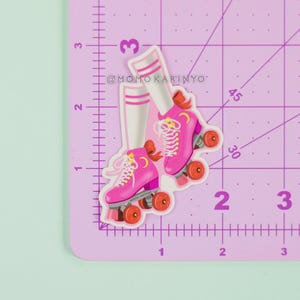 Può includere: Un adesivo con pattini a rotelle rosa con ruote arancioni e lacci bianchi, e calzini a righe bianche e rosa. L'adesivo è su un tappetino da taglio viola con misure in centimetri.