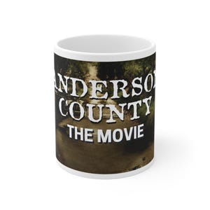 May include: White ceramic coffee mug with a wraparound design featuring the text "ANDERSON COUNTY THE MOVIE" in white block letters over a blurred outdoor scene with trees and water.