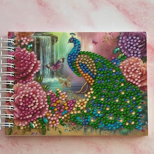 Könnte beinhalten: Ein spiralgebundenes Notizbuch mit einem lebendigen Pfauendesign. Der Einband zeigt einen Pfau mit einem juwelenartigen Körper, umgeben von Blumen, Schmetterlingen und einer Wasserfallszene. Die Farbpalette umfasst Blau-, Grün-, Rosa- und Lilatöne.