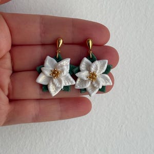 Può includere: Un paio di orecchini a forma di stella di Natale bianchi con dettagli dorati. Gli orecchini presentano petali dettagliati e foglie verdi, con un perno dorato. Un accessorio festivo.