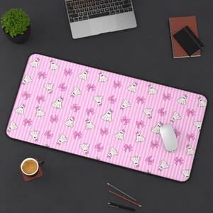 May include: Pink mousepad with a repeating pattern of white cartoon ghosts and pink bows on a striped background. A computer mouse rests on the pad, with a laptop, notebook, and coffee cup nearby.