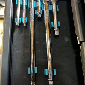 Könnte beinhalten: Eine Sammlung von Chrom-Steckschlüsseln und Verlängerungen, ordentlich in einer schwarzen Schublade mit blauen Haltern angeordnet. Die Werkzeuge variieren in Größe und Form, geeignet für Kfz- oder mechanische Arbeiten.