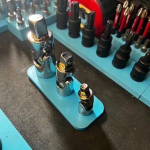 May include: A selection of chrome and black tools, including sockets and bits, arranged on a light blue tray. The tools are of varying sizes and shapes, suggesting a set for mechanical or repair work. Additional tools are visible in the background.