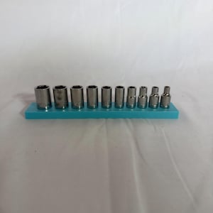 Könnte beinhalten: Ein Set aus zehn silberfarbenen Metall-Steckschlüsseleinsätzen, die in einer Reihe auf einer rechteckigen, hellblauen Kunststoff-Aufbewahrungsschiene angeordnet sind. Die Steckschlüssel variieren in der Größe und nehmen von links nach rechts im Durchmesser ab, wobei jedes Teil eine hohle zylindrische Form mit einer sechseckigen Öffnung an der Basis aufweist.