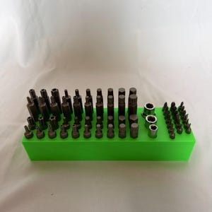 Könnte beinhalten: Ein hellgrüner, rechteckiger Aufbewahrungsblock mit einer Vielzahl von Metall-Werkzeugbits und drei silbernen Steckschlüsseln. Die Bits sind in Reihen angeordnet und weisen verschiedene Stern-, Sechskant- und Schraubendreherformen auf.