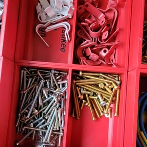 May include: A red plastic organizer tray with multiple compartments containing various hardware items including silver metal wall anchors, white plastic cable clips, red metal pipe clamps, gold screws, silver nails, brass bolts, and electrical wire.