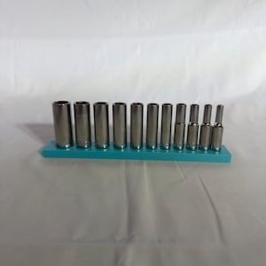 Könnte beinhalten: Ein Set aus zwölf silberfarbenen Metall-Steckschlüsseln, die nach Größe absteigend auf einer rechteckigen, türkisfarbenen Kunststoff-Aufbewahrungsschiene montiert sind. Jeder zylindrische Steckschlüssel hat eine hohle Mitte.