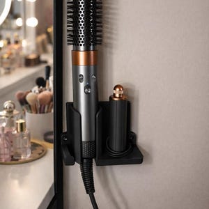 May include: A matte black wall-mounted storage holder for hair styling tools, featuring two distinct slots. One slot holds a grey and copper-toned hair dryer brush with a black power cord, while the other holds a separate black and copper attachment.