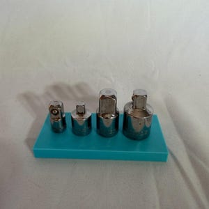 Könnte beinhalten: Ein Set aus vier silberfarbenen Metall-Steckschlüsseladaptern unterschiedlicher Größe, die in einer Reihe auf einem rechteckigen türkisfarbenen Kunststoffsockel angeordnet sind. Jeder Adapter hat ein quadratisches Ende und einen zylindrischen Körper.