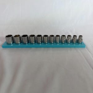 Könnte beinhalten: Ein Set aus zwölf silberfarbenen Metall-Steckschlüsseleinsätzen, die in einer Reihe auf einer rechteckigen hellblauen Kunststoff-Aufbewahrungsschiene angeordnet sind. Jeder Einsatz hat eine sechseckige Öffnung und eingravierte Größenangaben.