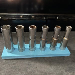 Könnte beinhalten: Ein Set aus sechs silberfarbenen Metall-Steckschlüsseln, die in einer Reihe auf einem hellblauen, rechteckigen Kunststoffhalter angeordnet sind. Jeder zylindrische Steckschlüssel hat ein hohles Inneres und eine metallische Oberfläche.