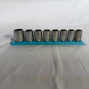 May include: A set of eight silver-coloured metal socket wrench attachments arranged in a single row on a rectangular teal plastic holder. Each cylindrical socket features a hollow centre and engraved size markings on the exterior surface.