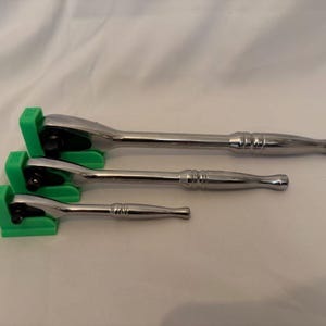 Könnte beinhalten: Drei silberfarbene Metallknarren in verschiedenen Größen, die jeweils in einer leuchtend grünen Kunststoffhalterung liegen. Die Knarren haben eine polierte Oberfläche mit Griff, Kopfmechanismus und Vierkantantrieb. Die Halterungen sind treppenförmig angeordnet.