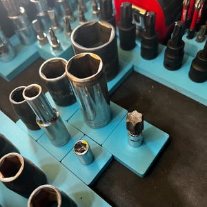 Puede incluir: Una colección de llaves de vaso y puntas metálicas, organizadas en bandejas de color azul claro. Se ven herramientas de varias formas y tamaños, incluyendo puntas hexagonales y en estrella. También hay una cinta métrica roja.