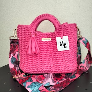 Peut inclure: Un sac en crochet rose vif fait à la main, avec un pompon et une poignée assortis. Le sac est doté d'une bandoulière à motifs floraux et d'une quincaillerie dorée. Une étiquette avec le texte "handmade" est attachée au sac.