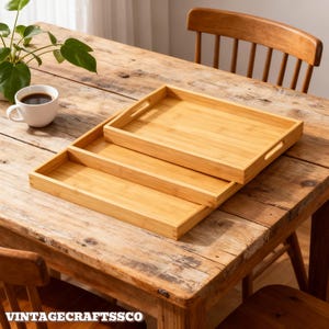May include: Three rectangular bamboo serving trays of varying sizes are stacked on a rustic wooden table. The trays have built-in handles. A white coffee cup and a plant are in the background. The text "VINTAGECRAFTSCO" is at the bottom.