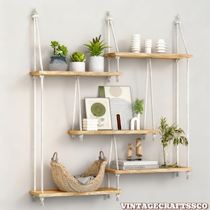 May include: A set of four wooden hanging shelves suspended by white rope. The shelves are adorned with potted plants, decorative vases, framed art, and a woven basket. The shelves are attached to the wall with metal hardware. The text "VINTAGECRAFTSCO" is at the bottom.