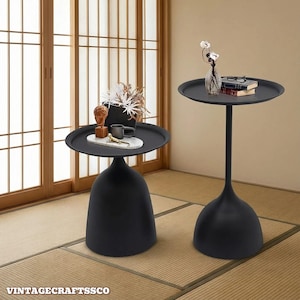May include: Two black accent tables with round tops and unique bell-shaped bases. One table is shorter and wider, while the other is taller and more slender. The tables are on a tatami mat floor, with a wooden door in the background.