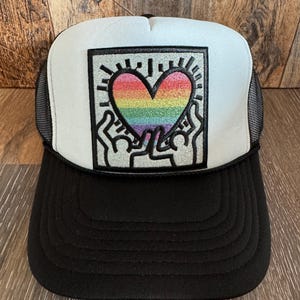 May include: Black and white trucker hat with a rainbow heart graphic. The heart is held by two silhouetted figures. The hat has a black brim and mesh back, with a white front panel.