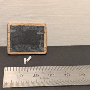 May include: A miniature rectangular chalkboard with a light brown wooden frame and two small pieces of white chalk. A metal ruler is positioned in the foreground, showing the chalkboard measures approximately 35 millimeters in width.