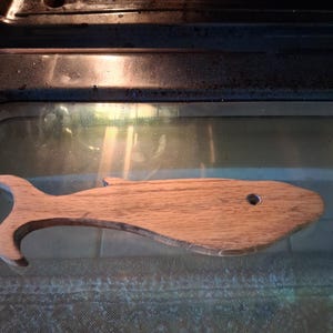May include: Wooden whale-shaped kitchen utensil with a hole and a curved tail. The light brown wood grain is visible. The utensil is inside an oven.