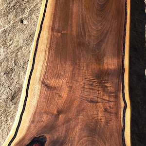 May include: A handcrafted wooden serving board with a natural edge and a handle. The board is made of dark brown wood with a black border and a red resin-filled knot. The board measures approximately 20 inches long.