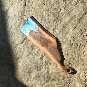 May include: A wooden serving board with a handle and a blue resin accent. The board is made of dark wood and has a natural grain pattern. The blue resin is at the top of the board, creating a decorative element.