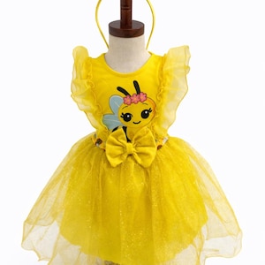 May include: A yellow bee-themed costume for a child, featuring a tutu skirt and a top with a bee graphic. The costume includes a headband with bee and flower accents. The dress is displayed on a mannequin.