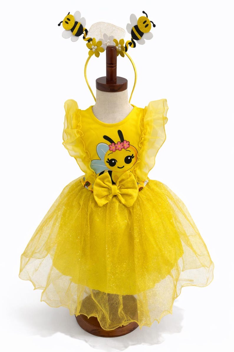 May include: A yellow bee-themed costume for a child, featuring a tutu skirt and a top with a bee graphic. The costume includes a headband with bee and flower accents. The dress is displayed on a mannequin.